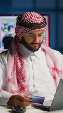 Dikey Arap adam online alışveriş yapıyor, kredi kartı bilgilerini bilgisayarda dikkatlice yazıyor. Müslüman kişi ödemeyi web sitesine yerleştirirken rahat ve sıcak bir evde geceleri