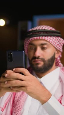 Oturma odasında gülümseyen Arap adam akıllı telefondan mesaj yazıyor. Orta Doğulu bir insan elinde cep telefonu tutuyor, arkadaşlarıyla internet mesajlaşması üzerinden sohbet etmekten keyif alıyor.