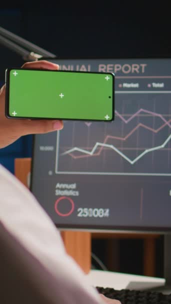 纵横视频男人拿着绿色屏幕手机 一边看电脑上的商业年报图表 自雇人士 可在家中使用彩色按键手机进行手工操作 — 图库视频影像