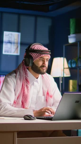 阿拉伯男子在家里客厅里带着无线耳机 在笔记本电脑上观看教育视频的垂直视频 中东人听播客 享受轻松的闲暇时间 — 图库视频影像