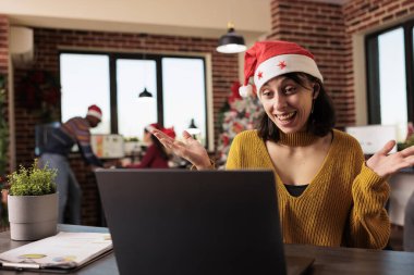 Gülümseyen kadın çalışan, dizüstü bilgisayardaki telekonferans uygulamasını kullanarak Noel süslemeli ofiste projenin son teslim tarihini tartışıyor. Noel Baba şapkalı neşeli proje yöneticisi video çağrısında iş arkadaşlarını selamlıyor