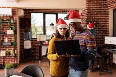 Şirket çalışanları birlikte çalışıyor, birlikte duruyor ve Noel 'de şirket ortamında dizüstü bilgisayarlarını tutuyorlar. Noel Baba şapkalı bir adam ve kadın Noel arifesinde projede çalışıyor.