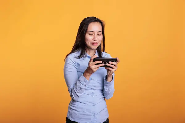 Playful gamer playing video games on mobile phone, trying to win gaming competition. Filipino woman having fun with online gameplay as leisure activity, standing in studio over yellow background