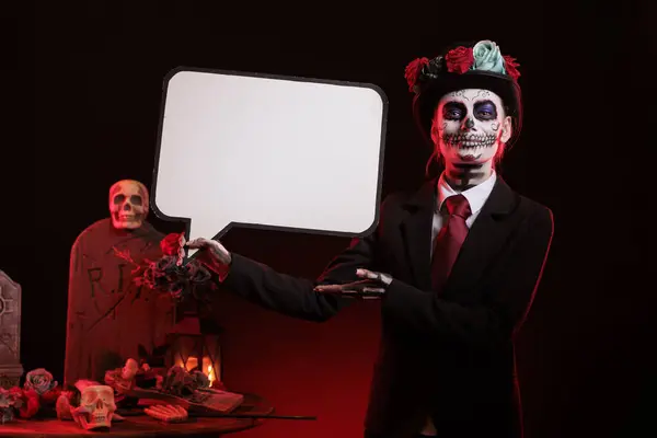 stock image Woman dressed in la cavalera catrina holding speech bubble with carnival make up and traditional day of the dead costume. Advertising isolated mockup cardboard and looking like santa muerte.