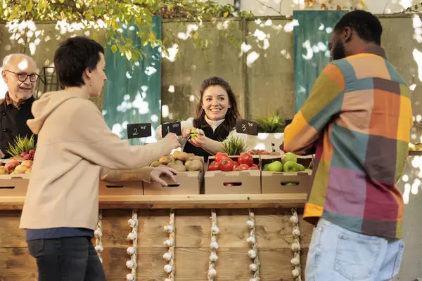 Young diverse family couple tasting different apple varieties while buying fresh organic fruits and vegetables at local farmers market. Friendly farm produce stand owner offering free samples