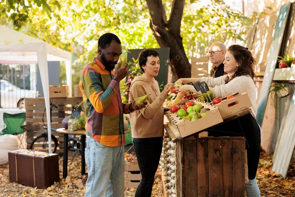 Diverse family tasting fresh organic products and doing shopping at outdoor farmers market stall. Friendly farmer vendor standing behind farm stand offering customers to taste bio apples.