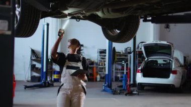 Tamir atölyesindeki arabanın altındaki titiz teknisyen bakım sırasında hasarı kontrol etmek için iş lambasını kullanıyor. Garaj işçisi, araç incelemek için profesyonel araç kullanıyor.