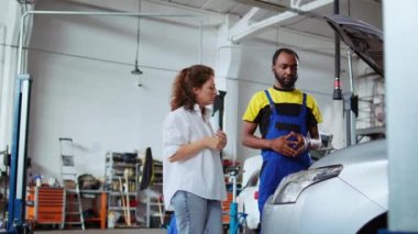 Araba servisinde deneyimli bir tamirci, müşteriye arabasının düzgün çalışabilmesi için nelerin değiştirilmesi gerektiğini gösteriyor. Asansöre bindirilmiş profesyonel teftiş aracı müşteriyle sohbet ediyor.
