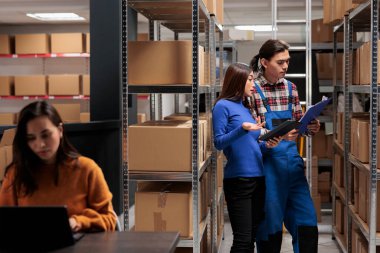 Depo yöneticileri sipariş listelerini pano ve dijital tablet üzerinde karşılaştırıyorlar. Asyalı çalışanlar karton kutularla dolu rafın yanında durup envanter raporlarını tartışıyorlar.