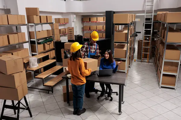 Warehouse package handlers doing inventory control while coworking with logistics manager. Asian storehouse employees scanning cardboard box and creating invoice on laptop