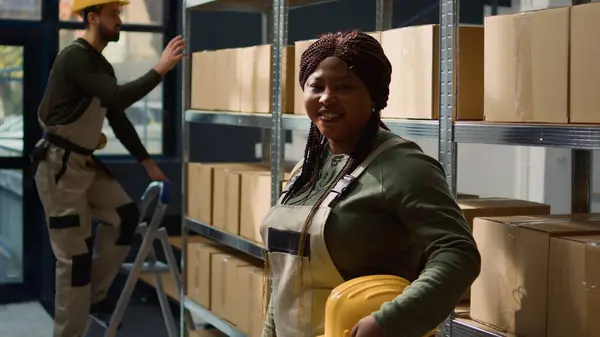 出荷する準備ができている段ボール箱の包装された商品で満たされた棚の前に立っている陽気なアフリカのアメリカの倉庫労働者の肖像画 専門の環境で働くこと幸せ — ストック写真