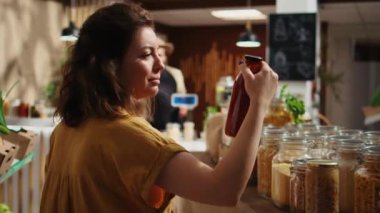 Sıfır atık süpermarketindeki mutlu kadın kasaya zımba taşımak için kağıt torba kullanıyor. Çevredeki bakkalda yaşayan yeşil müşteri yemek malzemeleri için alışveriş yapıyor.