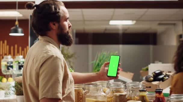 ゼロ廃棄物のスーパーマーケットで緑色のスマートフォンを使用して 健康的なレシピの成分をチェックします 地元の食料品店のクライアントは 有機野菜の買い物中にクロマキーフォンを使用しています — ストック動画