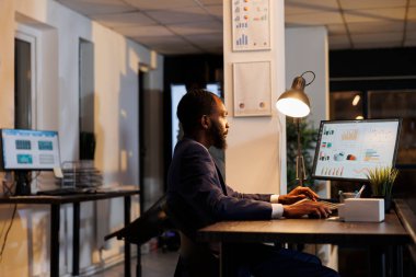 Afro-Amerikan işçi, pazarlama istatistiklerini analiz ediyor bilgisayardaki şirket stratejisini analiz ediyor, başlangıç ofisinde masa başında duruyor. Finans raporunda fazla mesai yapan girişimci. İş konsepti