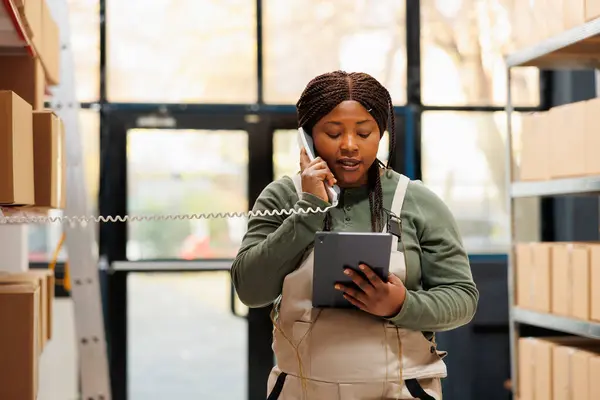 Manager answering landline phone call in warehouse, talking with remote supervisor about products quality control. African american worker wearing overall while checking logistics on tablet