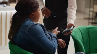 Müşteri, otel lobisinde boş oda beklerken servis edilen kahve fincanının parasını ödemek için KFK 'ye ödeme yapıyor. Afro-Amerikalı kadın belboyla lüks hizmetler hakkında konuşuyor. El kamerasıyla..