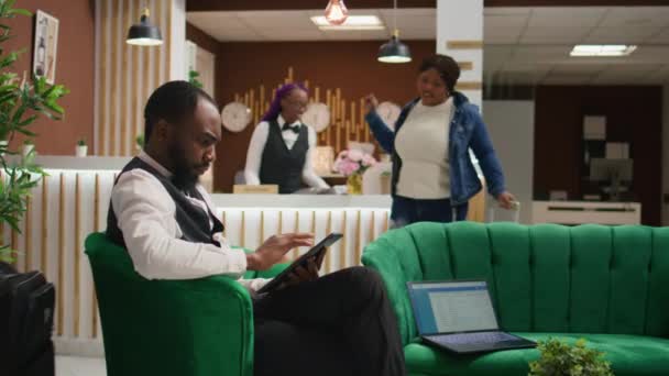 アフリカ系アメリカ人スタッフがロビーのタブレットでオンライン予約を行い 豪華なホテルで働き 到着時にゲストに優れたコンシェルジュサービスを提供しています ユニフォームマネージメントルームのベルボーイ — ストック動画