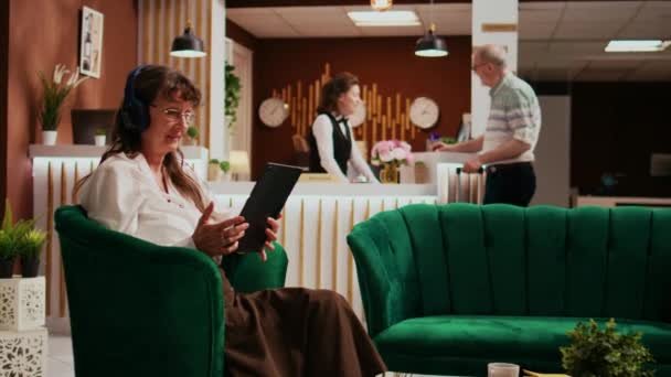ロビーでタブレットを使用して引退した旅行者は 宿泊施設を見るのを待っている間 ストリーミングサービスで映画を見ます ガジェットとヘッドフォンで現代技術を楽しんでいる高齢女性 — ストック動画
