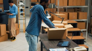 African american man putting products in cardboard boxes, preparing order for shipment. Young person doing quality control to ship merchandise to customers from storage room space.