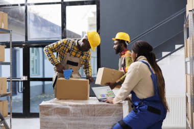 Afro-Amerikan depo yöneticileri müşteri siparişini hazırlıyor. Tüm siyah depo çalışanları karton kutuları yapışkan bantla paketliyor ve dizüstü bilgisayardaki mal listesini kontrol ediyor.