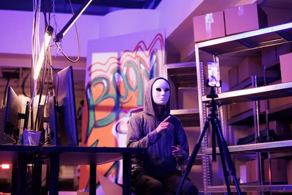 Hacker in anonymous mask threatening online while streaming live in abandoned warehouse. Internet scammer recording video with ransom on smartphone in hideout place at night