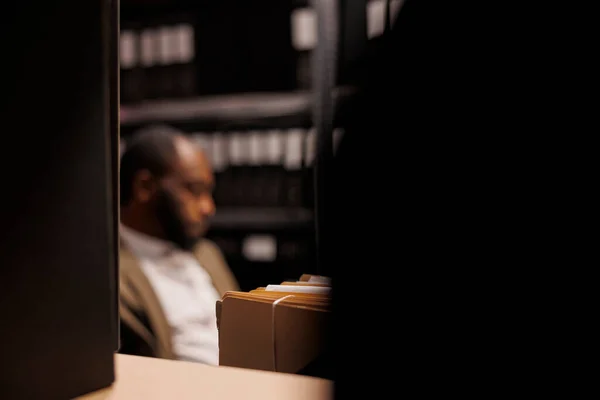 Shelf with criminal case files and policeman in dark police office. Blurred african american private investigator working overtime, solving crime at night time selective focus