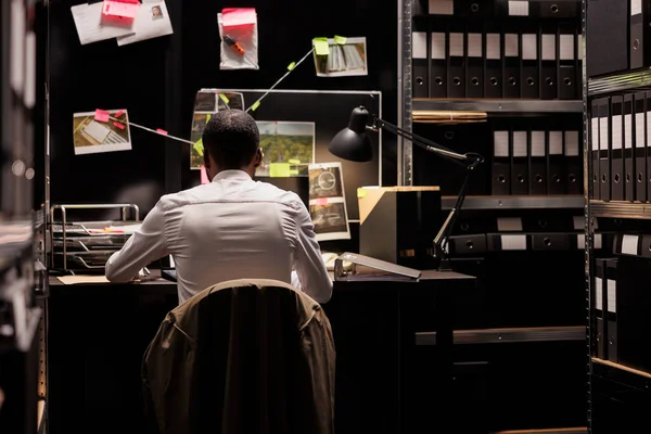 Detective sitting at desk and analyzing evidence scheme hanging on wall. Police law enforcement professional solving crime, studying clues and crime scene photos on board at night time