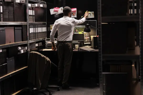 Private investigator putting crime case photo on detective board, analyzing clues. African american cop looking at proofs photographs and evidence with sticky notes hanging on wall