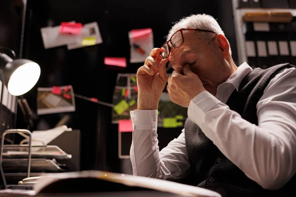 Tired elderly investigator working overtime in arhive room, analyzing criminal case evidence. Overworked private detective sitting at desk, checking mysterious suspect profile. Investigation concept
