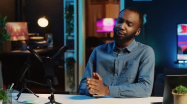 İnternetteki yayın platformları için video çekmek için cep telefonu kamerasını kullanarak loş ışıklandırılmış ev stüdyosu yaratıcısı. Etkilenen kişi, görüntüleri kaydetmek için cep telefonunu kullanarak seyirciyi selamlıyor