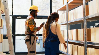 Team of employees scanning merchandise in depot, using scanner and tablet to check barcodes for stock logistics. Man and woman working on inventory, cargo pallets on racks and shelves. Handheld shot.