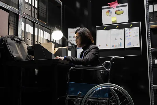 Asian accountant in wheelchair checking administrative analytical data in cabinet room office surrounded by accountancy statistic reports. Businesswoman with disability in file storage room