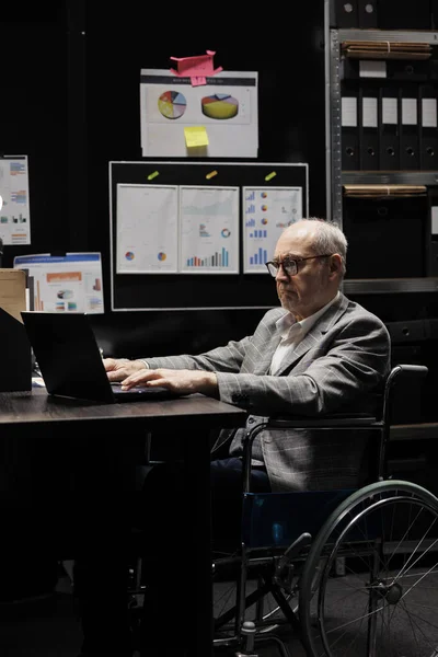 Senior businessman executive in wheelchair at office desk checking quarterly company results analysis data on laptop. Accountancy workplace filled with paperwork folders on cabinet shelves