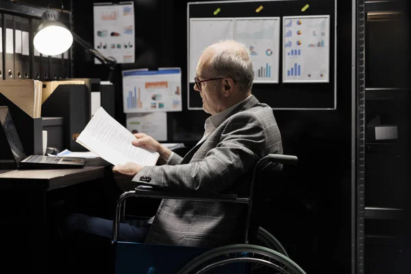 Professional accountant in wheelchair checking administrative paperwork in dark file room. Businessman in bureaucratic file cabinet office filled with invoice folders and flowcharts