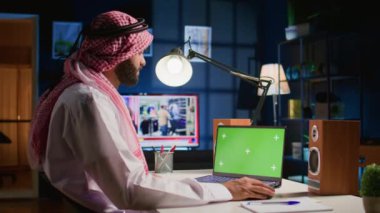 Orta Doğulu adam yeşil ekran dizüstü bilgisayarında, sosyal medya paylaşımlarını kontrol ediyor. Apartman dairesinde arka plan gürültüsü olarak televizyonu olan bir cihazdan internet sitelerine bakan serbest çalışan bir adam