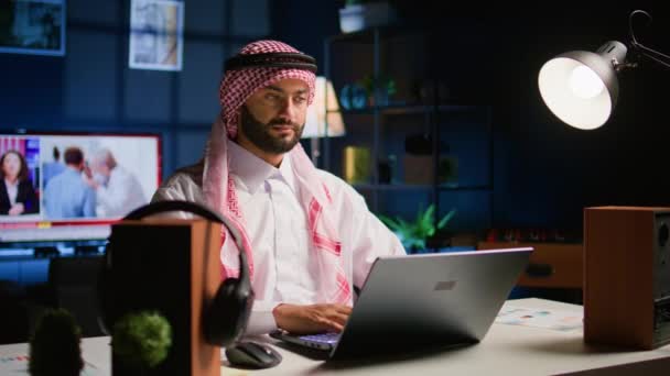 在时髦的公寓里愉快地回复工作邮件的阿拉伯电信工作者 快乐的穆斯林员工远程工作 在笔记本电脑上输入数据 打开电视作为背景噪音 手持相机拍照 — 图库视频影像