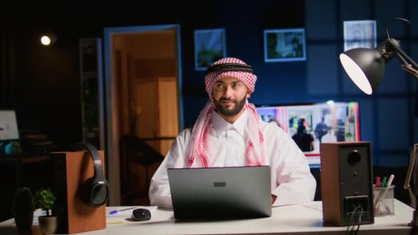 笑顔のアラブのテレワーカーはスタイリッシュなアパートで仕事の電子メールに答え すぐに仕事を終えることに満足しています 穏やかなイスラム教徒の従業員はリモートで働き バックグラウンドノイズとして開かれたテレビでラップトップ上のデータを入力します — ストック動画