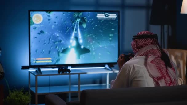 古典的なアーケードアクションビデオゲームをプレイするアラビアのゲーマーは 宇宙デブリを飛行します シングルプレイヤーゲームでミッションを解決するためにハイテクゲームコンソールを使用して自宅でレジャー時間を楽しむムスリム男性 — ストック動画