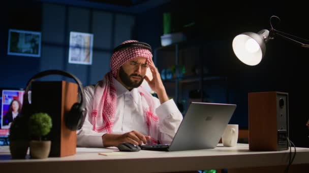 自宅のオフィスで働く片頭痛に苦しむ伝統的な服装の疲れたアラブのビジネスマン リビングルームのストレスを受けた中東の労働者は 期限に近づくことから頭痛を得る電子メールに答えます — ストック動画