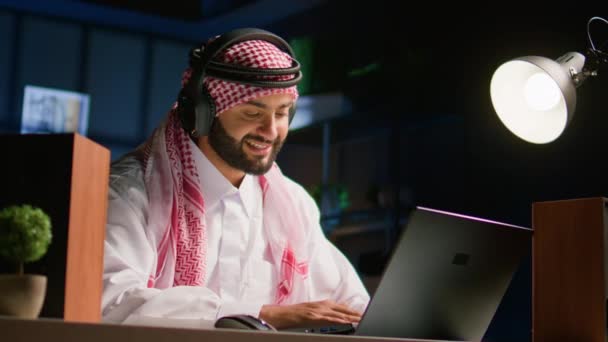 在家里客厅里拿着无线耳机 在笔记本电脑上打字的阿拉伯男人 穆斯林人听音乐 在笔记本上与朋友聊天 享受轻松的闲暇时光 — 图库视频影像