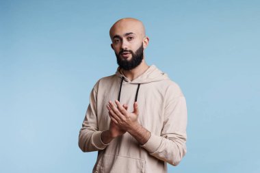 Arap adam ayakta durup kameraya tarafsız bir ifadeyle bakarken alkışlıyor. Kel sakallı genç adam kollarıyla alkışlıyor, stüdyo portresini kutluyor ve destekliyor.