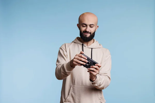 面白いアプリケーションソフトウェアを使用して モバイルゲームをプレイする若いアラブの男 青い背景のスタジオに立っている間スマートフォンでビデオゲームを楽しんでいる集中ゲーマー — ストック写真