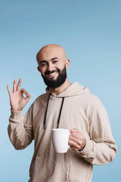 stock image Cheerful arab man holding coffee cup, smiling and showing ok gesture with fingers studio portrait. Carefree person drinking hot tea from white mug and looking at camera with approval sign