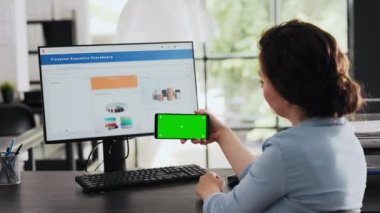 İş kadını elinde mobil cihaz, ofis masasında izole edilmiş kromakey ekran tutarken yeşil ekrana bakıyor. Çalışan telefondaki boş telif alanı yazılımını kontrol ediyor. El kamerasıyla..