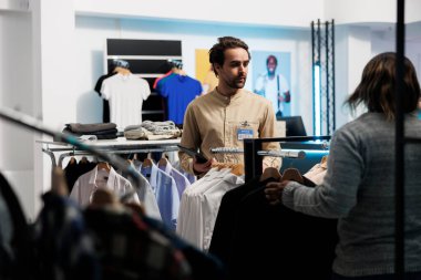 Alışveriş merkezi çalışanı, iş arkadaşıyla konuşurken giyim stokunu yönetmek için tablet kullanıyor. Giysi mağazasında çalışan beyaz adam butikte çalışırken web sitesinde giysileri inceliyor.