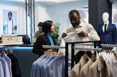 Afro-Amerikan giyim mağazası müşterisi askıda tişört tutuyor, stil tartışıyor ve danışmana uyuyor. Alışveriş merkezi galerisi Asyalı kadın asistanı müşteri tavsiyesi veriyor