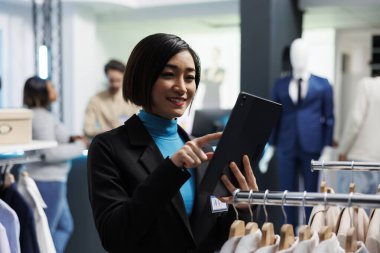 Gülümseyen Asyalı kadın perakende satış mağazasında çalışırken dijital tabletle giyim stoklarını analiz ediyor. Alışveriş merkezi çalışanı giysi ürünleri envanterini yönetiyor.