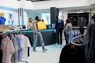 Alışveriş merkezi butiği tatmin olmuş bir müşteri giyim eşyalarıyla dolu çantalar tutuyor. Paketleri olan mutlu Afro-Amerikalı adam indirimli kıyafet aldıktan sonra eğleniyor.