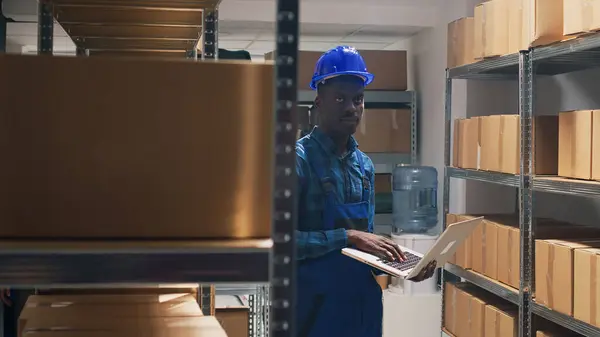 African american man using laptop in storage room, working on stock logistics to check cargo packed in cardboard boxes. Young adult holding pc and planning distribution, merchandise delivery.