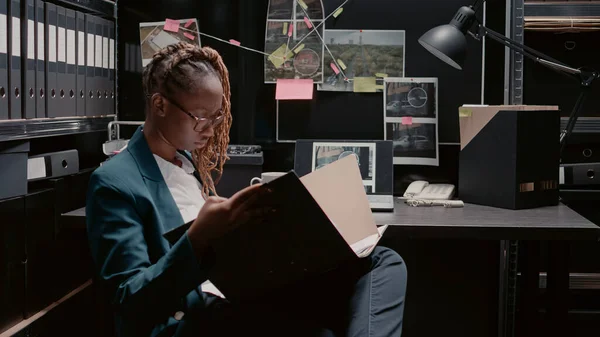 African american policewoman reviewing case files, working on criminal investigation with forensic evidence and suspect photos. Female inspector using detective board in law agency.